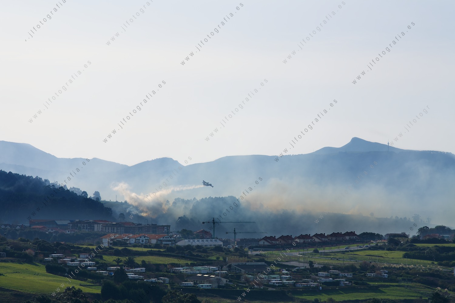 El pueblo de Berango, Getxo, con la humareda del incendio en los bosques próximos mientras el hidroavión descarga el agua