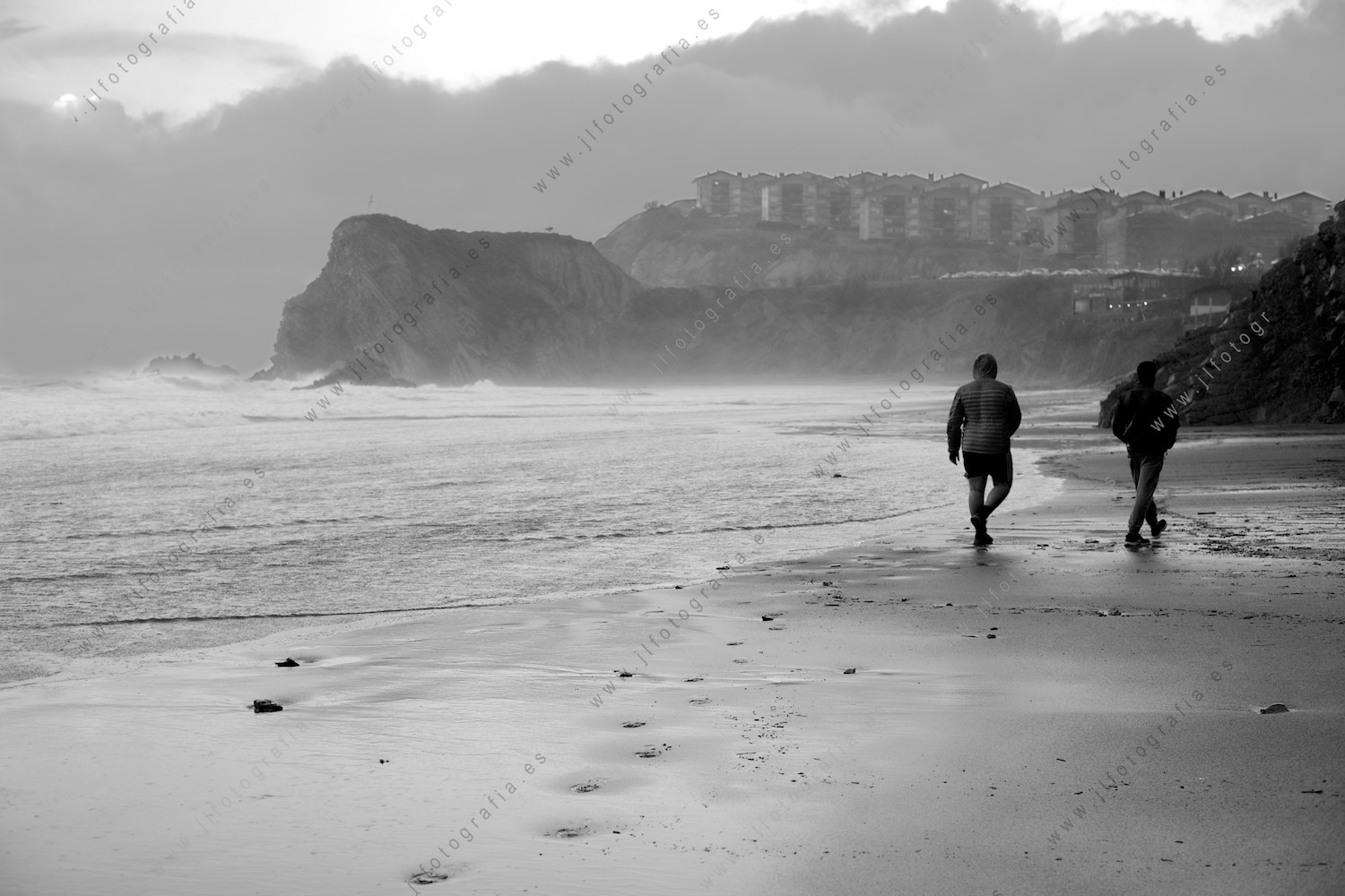 Dos hombres pasean por la playa de Sopelana, mientras las olas golpean fuerte en el fondo.