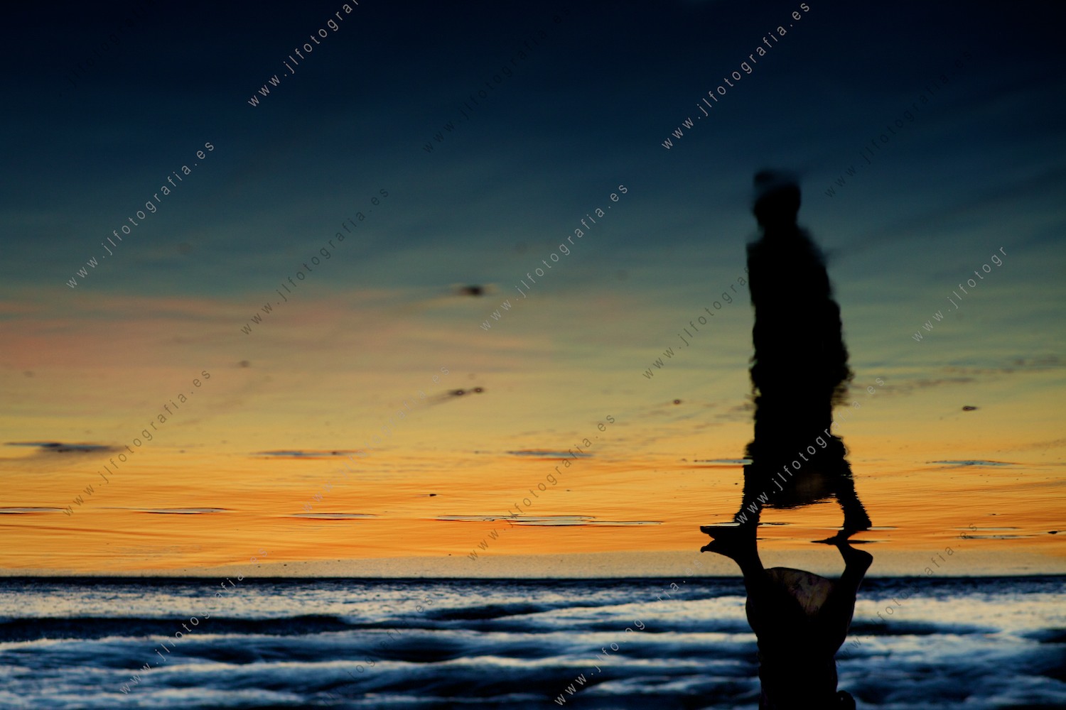 Los colores del crepúsculo, reflejados en la orilla de la playa juega con la silueta de una mujer que pasea.