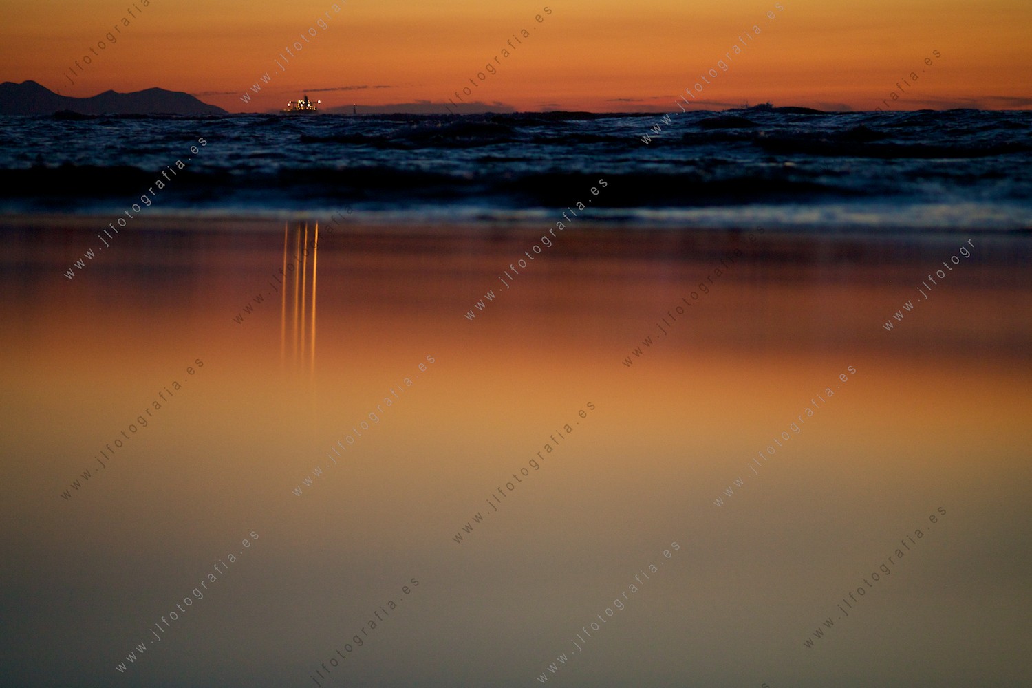 Un barco en el mar, refleja sus luces en el espejo que crea la arena empapada en la cálida luz crepuscular.