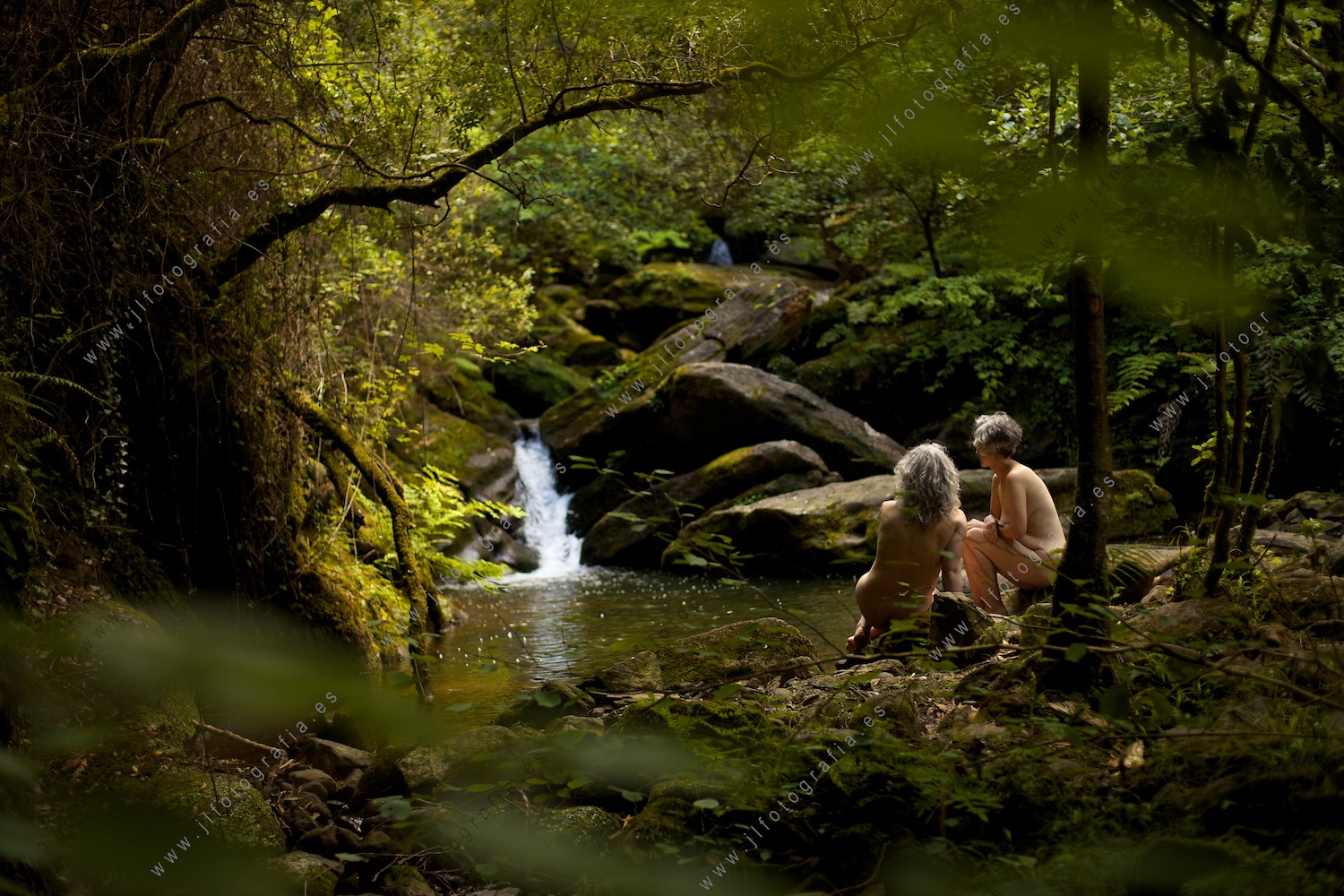 Dos mujeres desnudas sentadas en la orilla del río junto a una cascada.